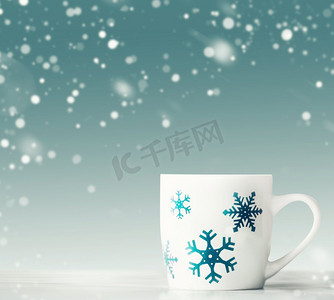 白色桌子上有蓝色雪花的白色马克杯，蓝色背景下有降雪，前景。寒假快乐卡片排版