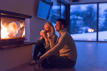 在寒冷的冬夜，年轻浪漫的情侣坐在壁炉前的地板上聊天喝茶