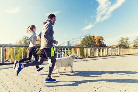运动女人摄影照片_健身，运动，人和慢跑的概念-快乐的夫妇与狗在户外跑步。一对幸福的夫妇带着狗在户外奔跑