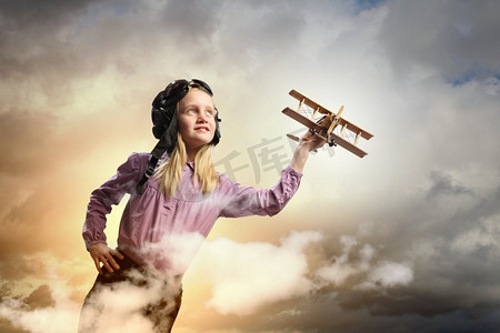 戴着飞行员S帽子的小女孩。戴着飞行员头盔的小女孩在云的背景下玩玩具飞机