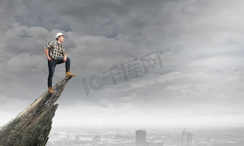 51巅峰钜惠摄影照片_年轻自信的建设者站在高高的城市上方的岩石边。攻克巅峰