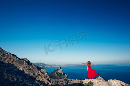 一位穿着红裙的年轻漂亮女子望着山海。蓝天背景下的大自然女孩。时尚写真