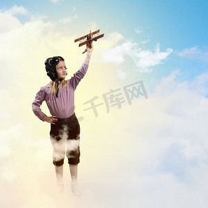 玩玩具的小女孩摄影照片_戴着飞行员头盔的小女孩在云的背景下玩玩具飞机
