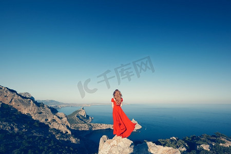 一位穿着红裙的年轻漂亮女子望着山海。蓝天背景下的大自然女孩。时尚写真