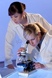 看显微镜摄影照片_一位美丽的女医学或科学研究人员在用她的显微镜，而她的东方同事在她身后看不到焦点。