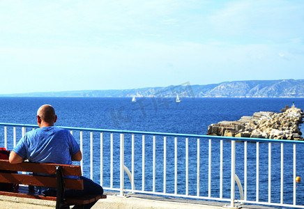 海滨长廊摄影照片_坐在马赛海滨长廊地中海沿岸长凳上的年轻男子旅行者。