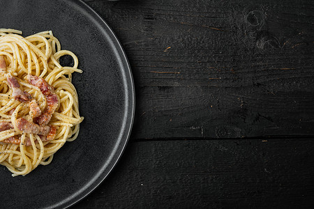 美食广告摄影照片_意大利面条 Carbonara 的菜肴，现代意大利面食配 guanciale，鸡蛋广告 pecorino romano 奶酪，黑色木质背景，顶视图平躺，带文本复制空间