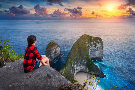 女人坐在悬崖上，在印度尼西亚巴厘岛努沙佩尼达岛的 Kelingking 海滩看日落。