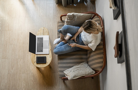 手机阅读器摄影照片_女人躺在沙发上使用智能手机，桌上放着笔记本电脑和电子阅读器。