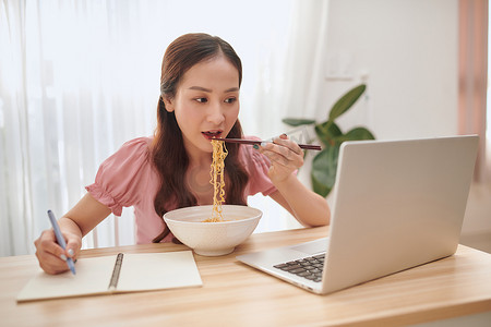 年轻的亚洲妇女在家里吃面条和用笔记本电脑工作。