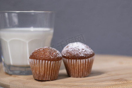 一杯牛奶杯摄影照片_在一杯牛奶旁边放两个小纸杯蛋糕或糖粉松饼。