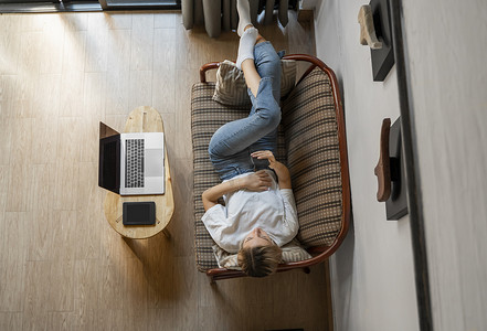 女人躺在沙发上使用智能手机，桌上放着笔记本电脑和电子阅读器。