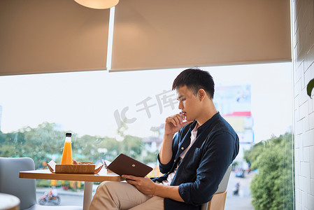 商人在咖啡厅窗边的笔记本上写字，专注于工作，自信的年轻人的侧视图在咖啡厅坐着写字时正在笔记本上工作，年龄 20-30 岁。