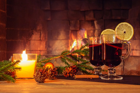 在燃烧的壁炉背景下，两杯酒、一支蜡烛、在木桌上装饰的冷杉树枝。