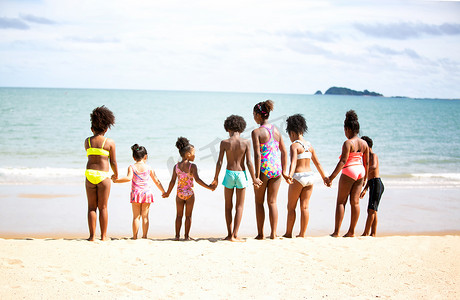 孩子们在沙滩上的沙滩上玩耍，一群孩子在夏天手牵着手在沙滩上排成一排，后视大海和蓝天