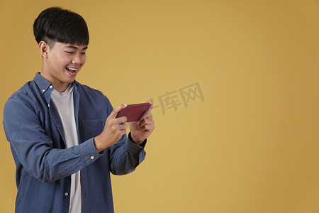 黄色背景中，兴奋的亚洲年轻男子穿着随意地在智能手机上玩电子游戏的肖像