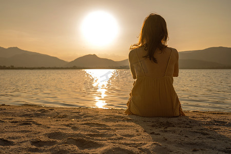 迷茫的摄影照片_无法辨认的孤独沉思的女人坐在海边的沙滩上，看着夕阳在水中的光反射和橙色棕褐色复古摄影效果。