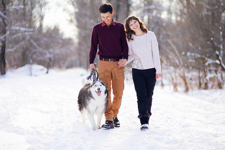 一对年轻夫妇带着他们的哈士奇狗在冬季公园里微笑和玩乐