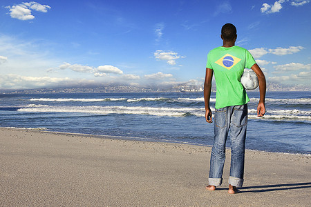 穿着巴西 T 恤站在沙滩上踢足球的男子的后视图