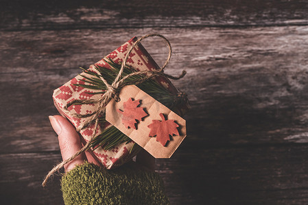 手拿着一份秘密的圣诞小礼物，里面有创意手工装饰质朴的 diy 礼物，用红色复古包装纸包裹着，用天然复古麻线和云杉​​树枝作为旧木背景上的装饰，送给特别的人