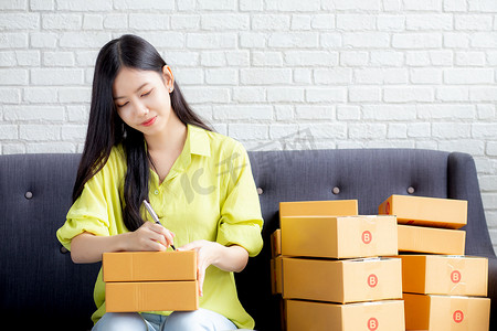 诚邀商家摄影照片_年轻的亚洲女性在家里的包裹箱上写地址，以便向客户、运输和物流、在线商家和卖家、企业主或中小企业、在线购物和电子商务订购送货单。