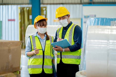 戴头盔摄影照片_两名带卫生面具的工厂工人一起讨论在仓库工作场所管理产品。