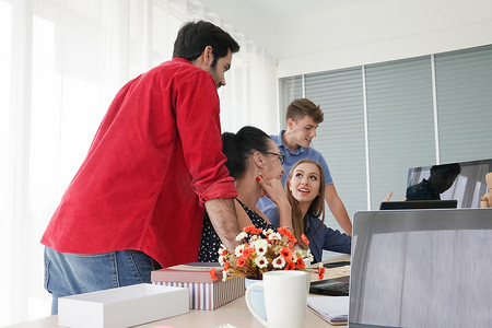 在办公室工作的商务人士使用台式电脑，一群穿着时髦休闲装的快乐商务人士看着笔记本电脑并打手势。