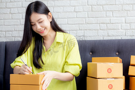 年轻的亚洲女性在家里的包裹箱上写地址，以便向客户、运输和物流、在线商家和卖家、企业主或中小企业、在线购物和电子商务订购送货单。