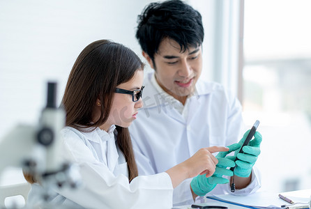年轻的白人科学家女童指着她的亚洲老师在实验室或课堂上解释实验分析的手机。
