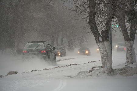 北方寒冷摄影照片_冬天，在暴风雪、暴风雪或降雪中，城市的恶劣天气下，积雪覆盖的道路上有汽车。北方的极端冬季天气条件。汽车驶过城市积雪覆盖的街道