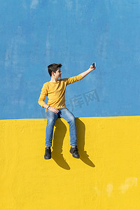 正面图是一个穿着休闲服的小男孩坐在黄色围栏上靠着蓝色墙壁，同时用智能手机自拍