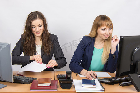 两个在办公室工作的年轻女性，一个有趣地拿了一张纸，第二个看着电脑屏幕