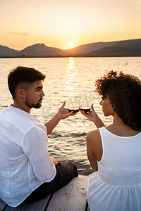 情侣浪漫场景摄影照片_多种族情侣在日落或黎明时坐在码头上的浪漫场景，用红酒敬酒，看着对方的眼睛 — 迷人的男人与她的西班牙裔女友结合 — 专注于眼镜