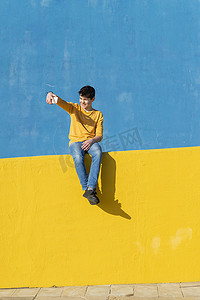正面图是一个穿着休闲服的小男孩坐在黄色围栏上靠着蓝色墙壁，同时用智能手机自拍