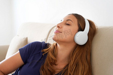 安静的千禧一代女孩躺在舒适的沙发上，戴着无线耳机，享受美妙的冥想音乐。
