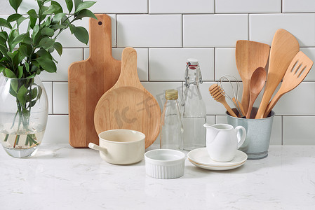水壶摄影照片_白色木墙上的简单质朴厨具：带木制炊具套装的粗糙陶瓷锅、成堆的陶瓷碗、水壶和木托盘。