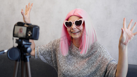 假发摄影照片_戴着粉红色假发的快乐女博主站在三脚架上的镜头前