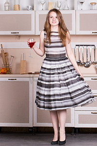 年轻漂亮的女人穿着裙子，在她的厨房里拿着一杯酒。