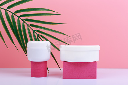 两个几何形状的奶油罐，用于在带棕榈叶的粉红色背景的白桌上展示产品