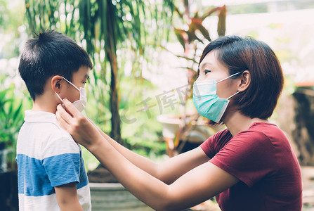 公共生活摄影照片_母亲在户外照顾带口罩的儿子以保护流感或 covid-19，妈妈戴上带儿童安全的医用口罩以保护公共场合大流行病的爆发，医学概念。