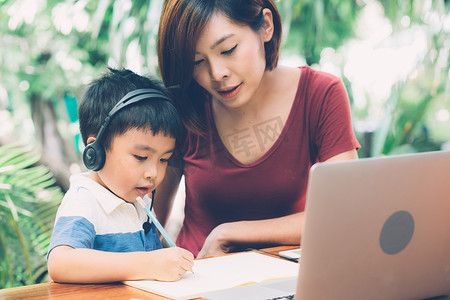 年轻的亚洲母子使用笔记本电脑在家学习和学习，男孩在笔记本上写作业并戴耳机，老师或妈妈支持孩子，教育理念。