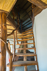 木楼摄影照片_在白墙附近，风格化的装饰性不寻常的木楼梯带有来自树枝的扶手，通向二楼，屋顶覆盖着稻草