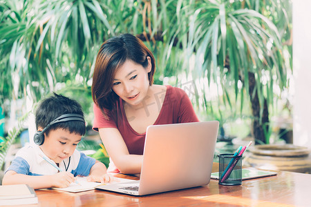 妈妈和孩子摄影照片_年轻的亚洲母子使用笔记本电脑在家学习和学习，男孩在笔记本上写作业并戴耳机，老师或妈妈支持孩子，教育理念。