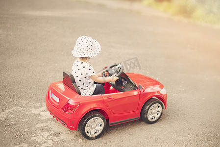 小女孩穿着白色黑头衬衫和帽子，在公园附近开着一辆红色玩具车。