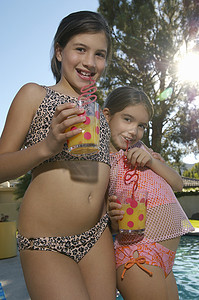 喝果汁的女孩摄影照片_穿着泳装的小女孩在泳池边用疯狂的吸管喝果汁的肖像