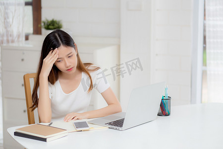 年轻的亚洲女性在笔记本电脑上工作时头痛，在家里的办公桌上有最后期限，失败的女商人筋疲力尽、压力大、担心、沮丧和失败。