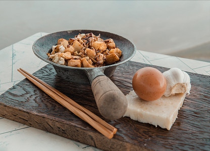 萝卜糕摄影照片_小蒸铁锅中的传统炒软萝卜糕或炒萝卜糕（chai tow kway），在白桌上用木筷子端上。