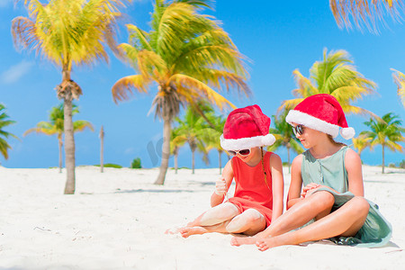 小孩圣诞老人摄影照片_可爱的小孩在圣诞海滩假期期间戴着圣诞老人的帽子玩得很开心。