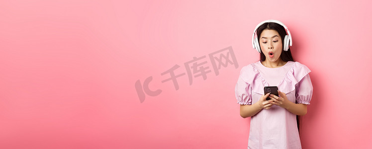 兴奋的亚洲女孩看着智能手机屏幕被逗乐了，说哇，用无线耳机听音乐，站在粉色背景下