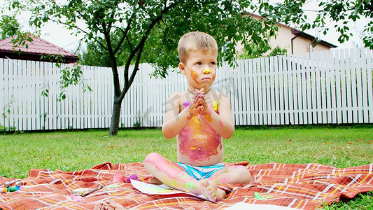 一个小孩，一个四岁的男孩在玩耍，用手指画画，装饰自己，在花园里，坐在毯子上，在草地上，草坪上，在夏天。